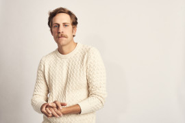 Male with a mustache wearing a cream sweater in front of a white background